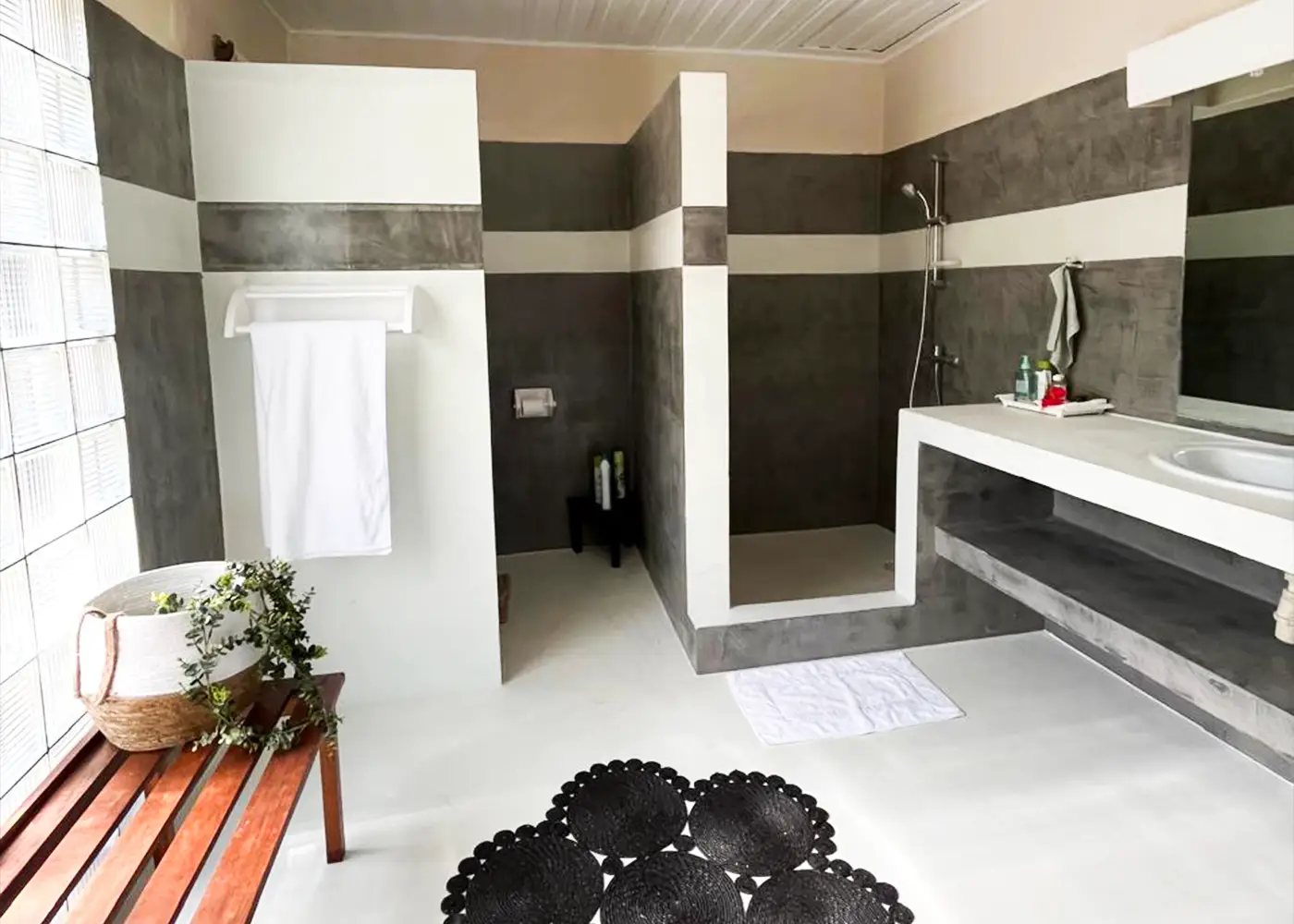Modern bathroom with gray and white tile, open shower, white towel, wooden bench, and plants.