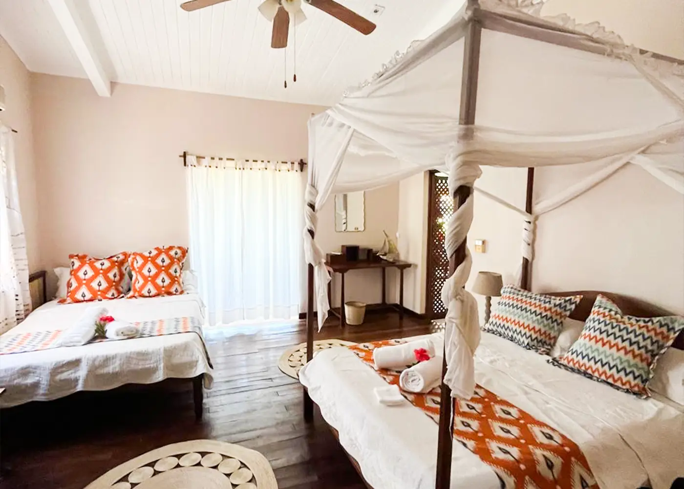 Bright bedroom with two beds, white linens, patterned pillows, a ceiling fan, and sheer curtains.