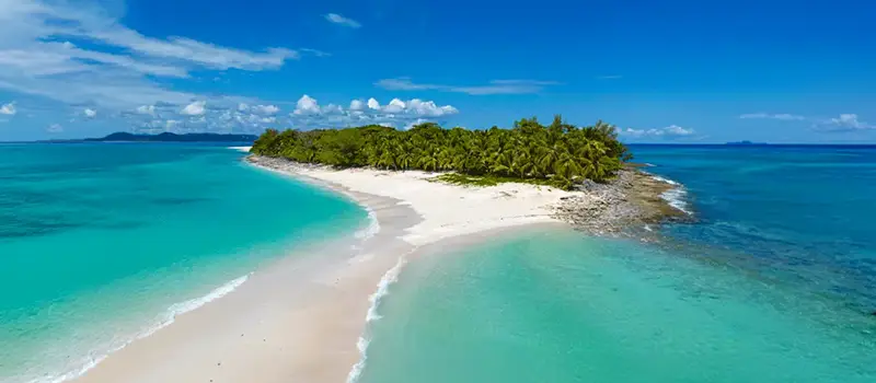 Aerial view of a small tropical island with white sand beach surrounded by clear turquoise ocean under blue sky.