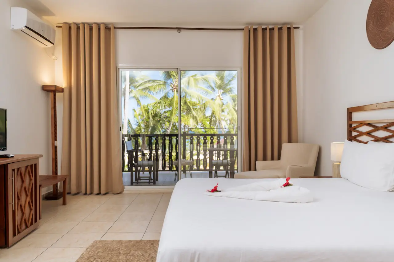 Hotel room with a large bed, beige curtains, armchair, and balcony showing palm trees outside.