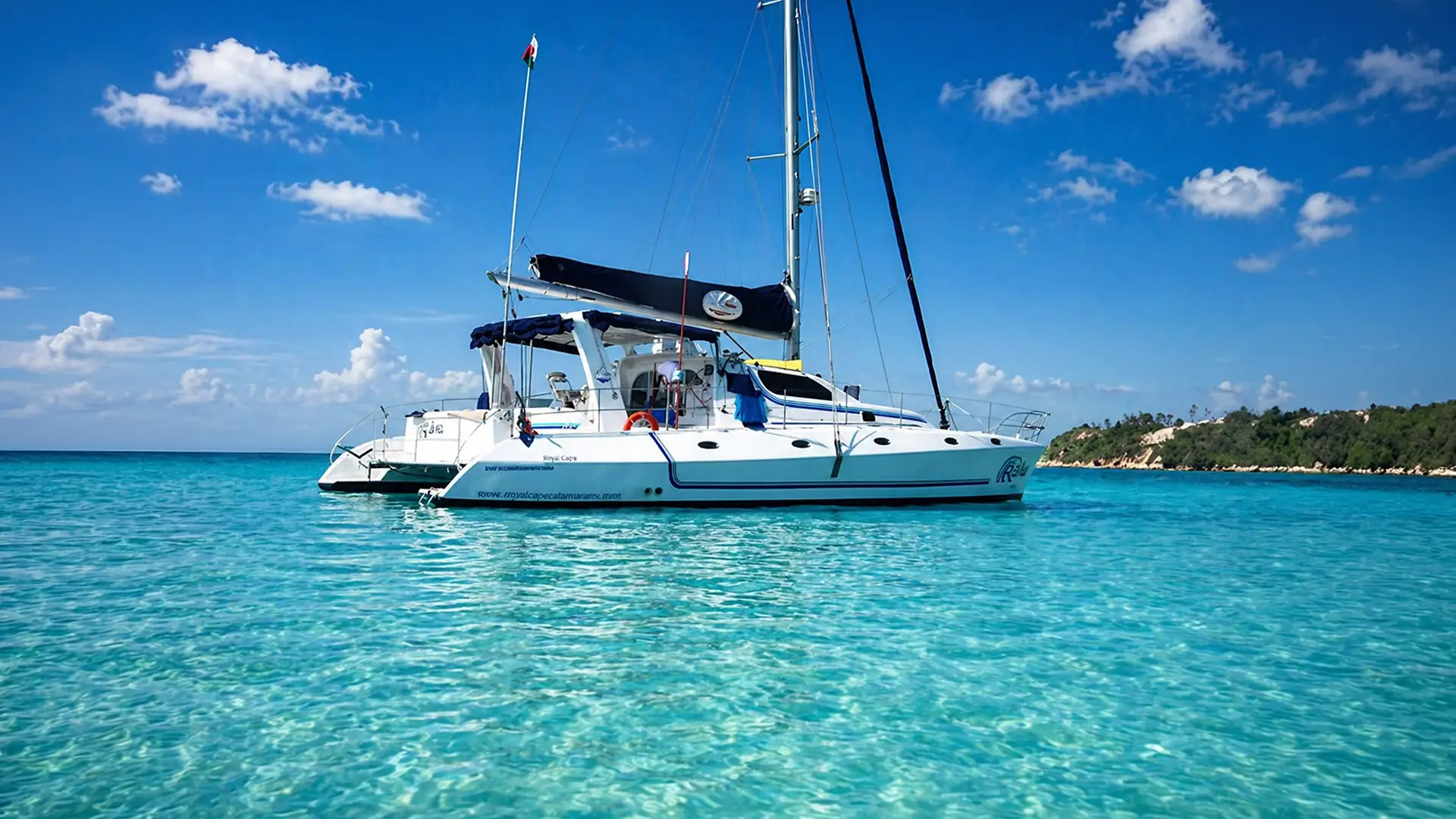 White catamaran sailboat anchored on clear turquoise water with a blue sky and distant green shoreline.