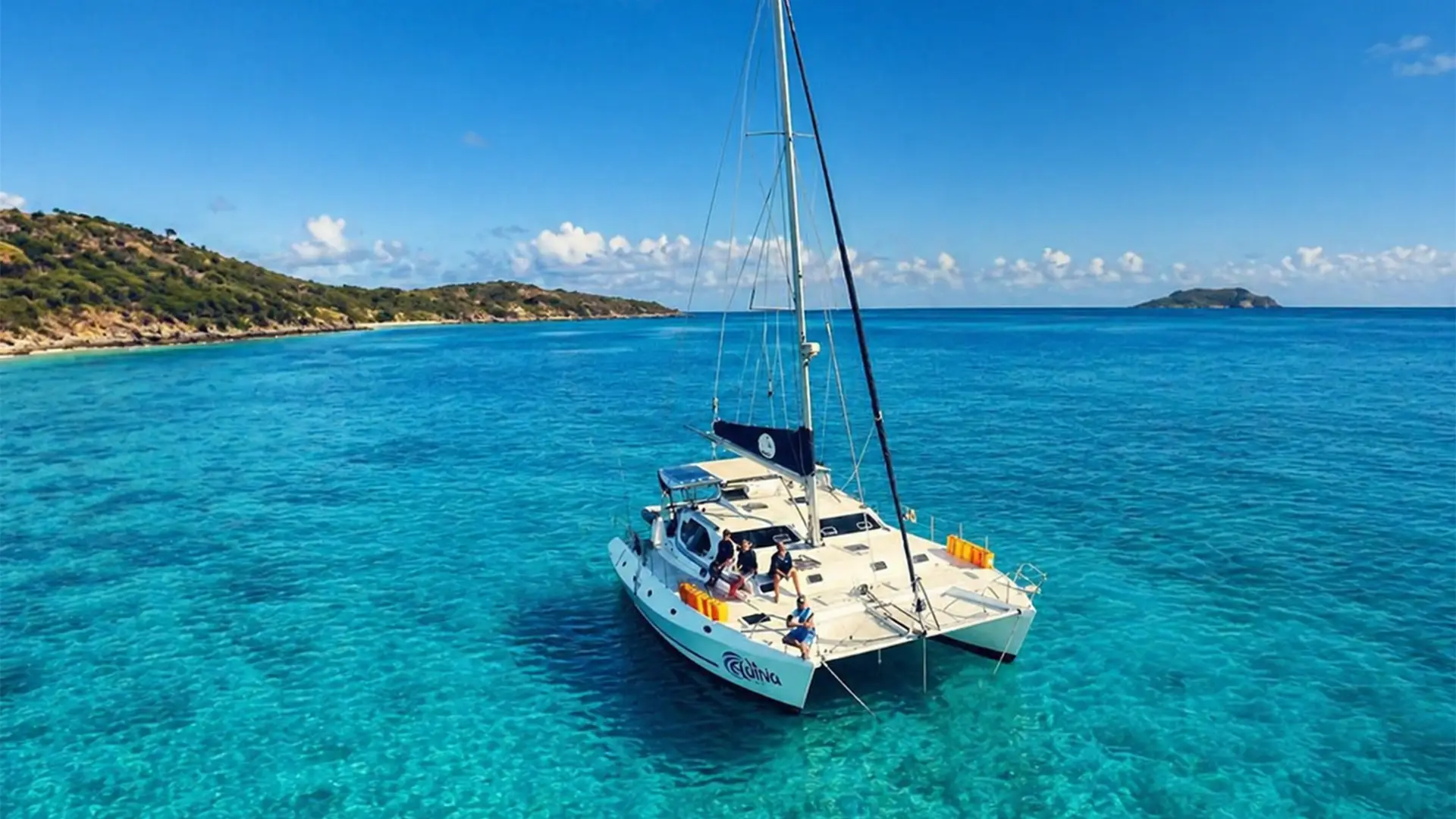 Catamaran yacht with people on deck floating on clear turquoise water near a green island.